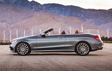 A silver Mercedes-Benz cabriolet is parked with the top down in front of a row of modern windmills.