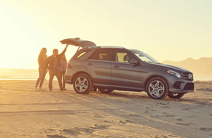 A group of people is closing the rear door of a Mercedes-Benz SUV parked on the shore. 