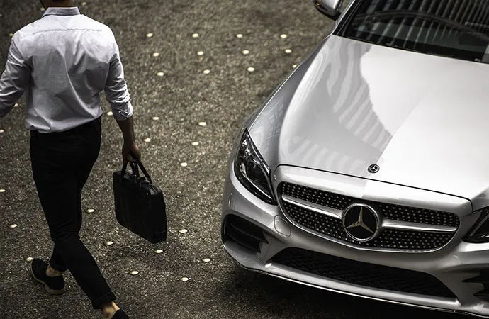 A man with a bag is walking away from a parked shiny silver Mercedes-Benz sedan.