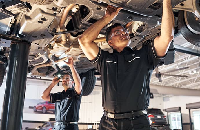 Two Mercedes-Benz service technicians work on the underside of a lifted vehicle.